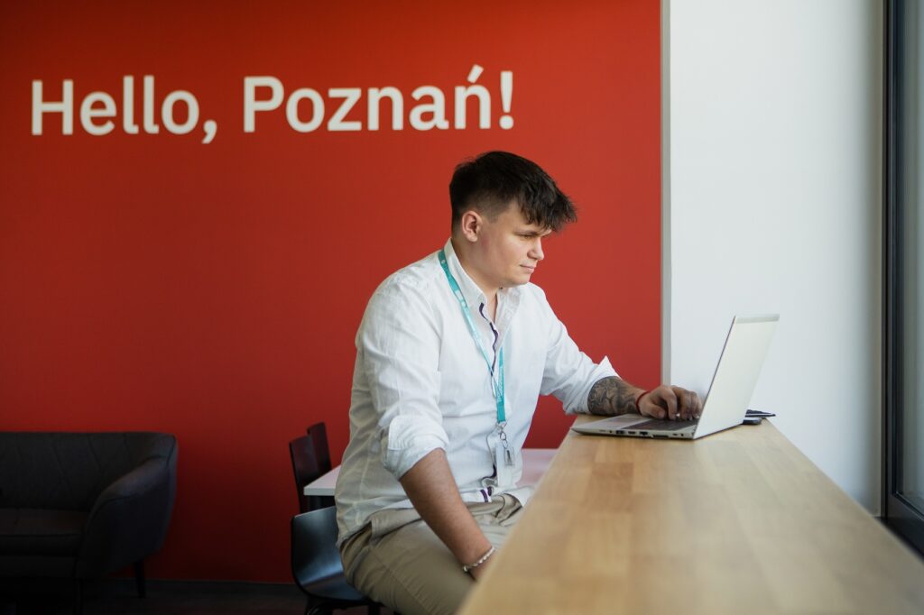 Pracownik Transcomu w białej koszuli siedzi przy biurku przed czerwoną ścianą z napisem "Hello, Poznań!".