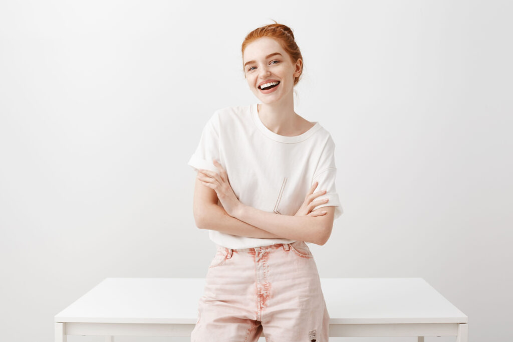 Młoda ruda dziewczyna uśmiecha się do kamery, siedząc na biurku na białym tle