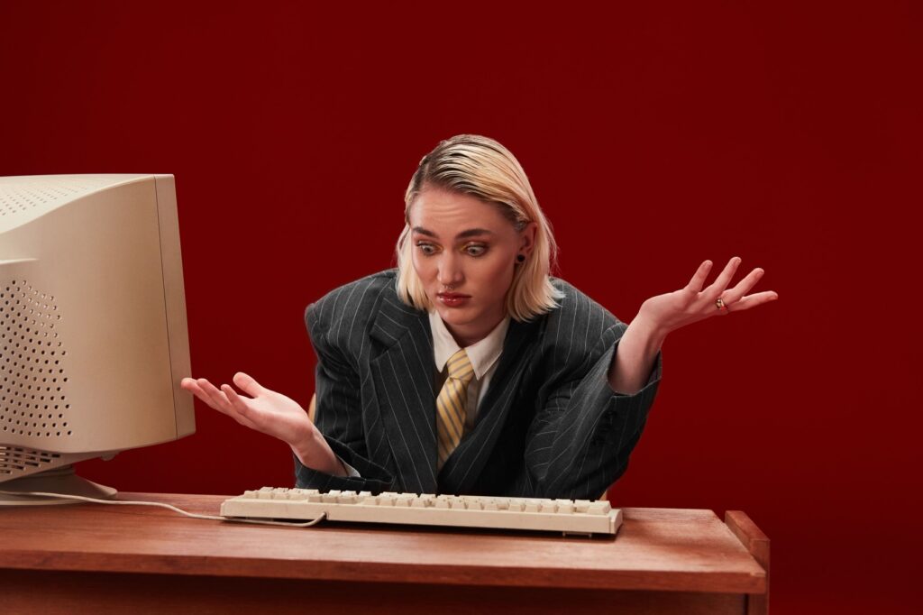 Młoda dziewczyna w garniturze rozkłada ręce ze zdziwieniem, siedząc przy biurku, na którym stoi retro komputer